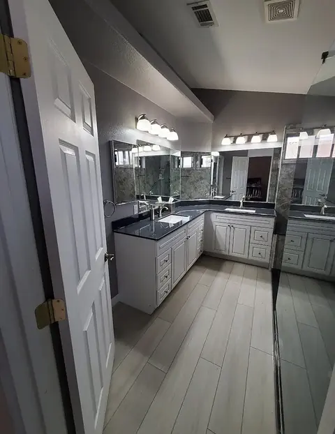 Double Vanity Master Bath — L-shaped double vanity with dark stone countertops, white cabinetry, framed mirrors and wood-look plank tile flooring, installed by Revive Bath Solutions in Athens, TX